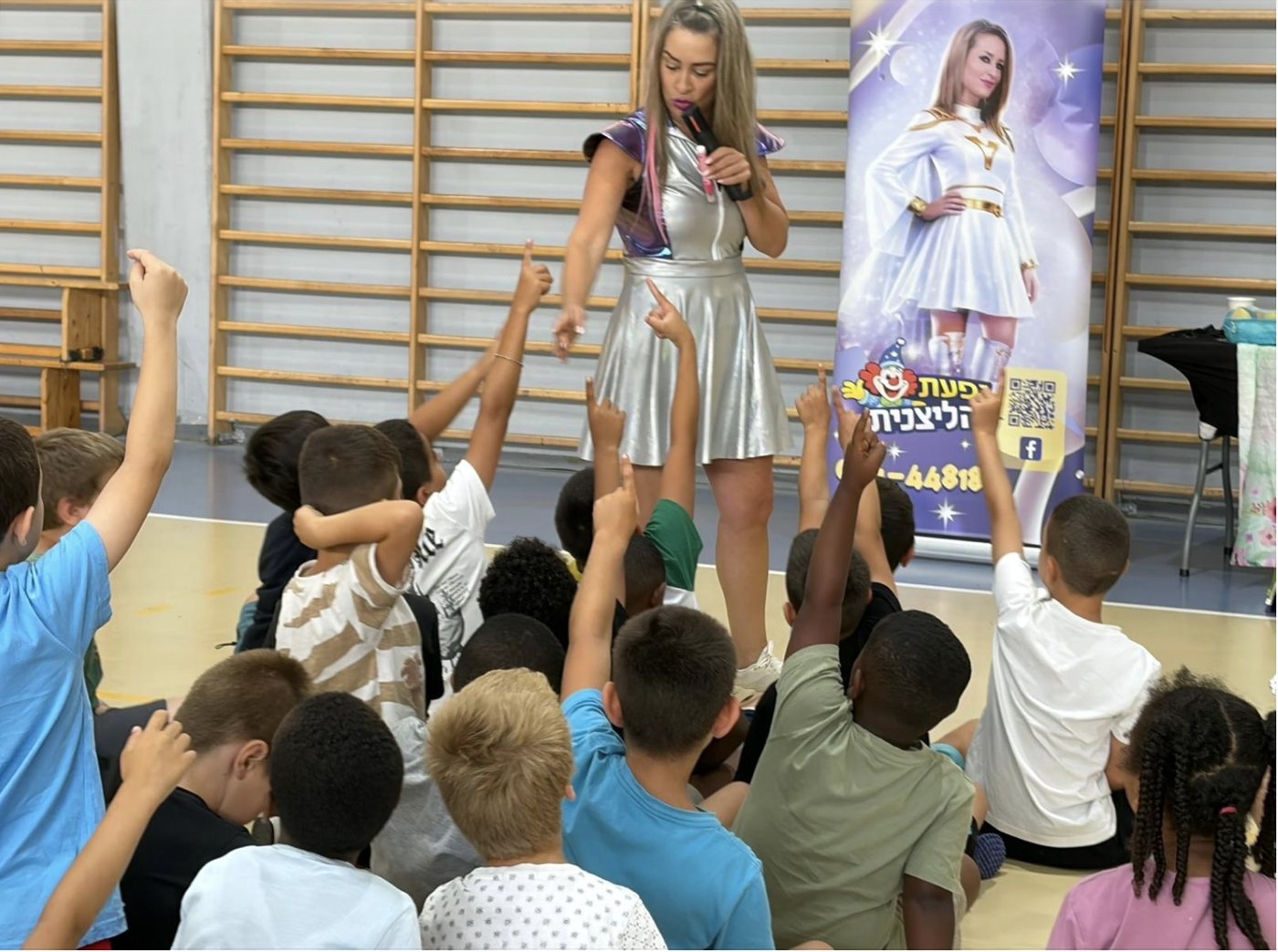 יפעת מפעילה במיקרופון קבוצת ילדים באולם ספורט, והילדים מרימים ידיים
